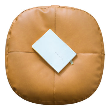 A round tan leather pouf or floor cushion with a white notebook resting on top, photographed from a top-down angle against a solid white background.