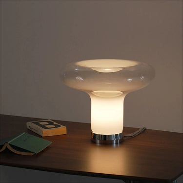 A modern mushroom-shaped table lamp with a glowing frosted glass body and a clear glass top, sitting on a dark wooden table next to a small book.