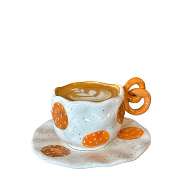 A ceramic coffee cup with orange and white pretzels pattern on the side, accompanied by a matching saucer.