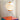 A modern table lamp with a ribbed white paper lantern shade, orange accents, and three black metal legs, shown on a colorful translucent pedestal against a white radiator background.