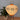 A Japanese-style paper lantern table lamp with a warm glowing beige body, black geometric cutout patterns, and orange-brown trim, displayed on a wooden surface next to a potted green plant against a gray wall.