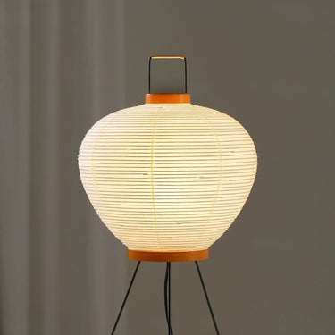 A Japanese-style paper lantern table lamp glowing with warm light, featuring a ribbed white paper shade, orange-brown wooden accents, and a black metal stand against a neutral gray background.