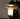 A person holding a modern mushroom-shaped table lamp with a clear glass shade and a glowing white illuminated base on a silver metal base, set against a plain gray wall.