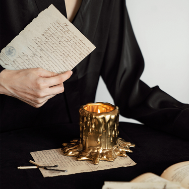 A gold-colored candle with a resin container, sitting on a piece of paper, with a person holding a book in the background.