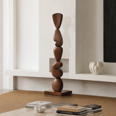 A tall, abstract wooden sculpture with stacked organic shapes on a rectangular base, displayed in a minimalist interior setting with books and a white vase on a shelf.