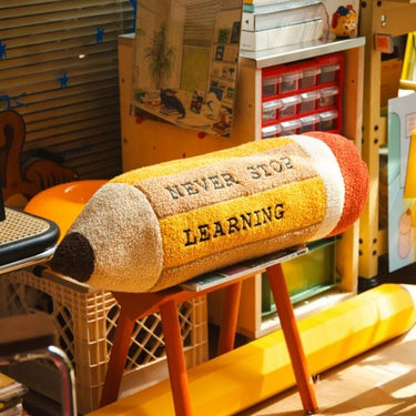 A plush pencil-shaped cushion with the words 'NEVER STOP LEARNING' embroidered on it, resting on a wooden stool in a colorful classroom or playroom setting.