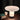 A mushroom-shaped table lamp with a glowing white frosted shade and cylindrical base, placed on a wooden table next to a small mug and books against a dark curtain background.