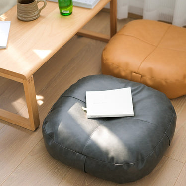 Two round floor poufs, one dark gray and one tan, on a light wood floor next to a wooden table with a mug and book.